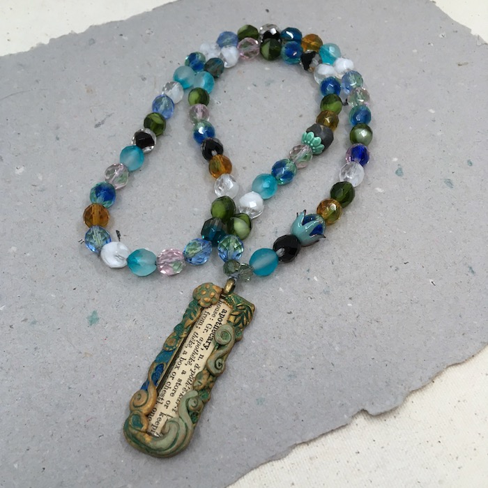 Pendant made of dictionary text surrounded by green, gold and blue pattern work with a necklace of green, blue, white and gold Czech glass beads. 